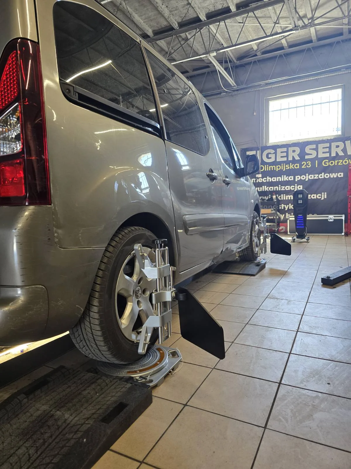 Citroën Berlingo na stanowisku geometrii kół 3D – obejma z targetem pomiarowym FCAR na tylnym kole, w tle baner Tiger Serwis Olimpijska 23